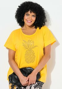 Camisa amarilla de manga corta con un gráfico de piña de strass. Combinada con pantalones estampados y un clutch negro. Textura de algodón suave.