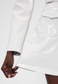 Belo, sijoče oblačilo iz lakiranega usnja z manšetnim rokavom, ki prikazuje razširjen kroj krila in subtilen detajl žepa.