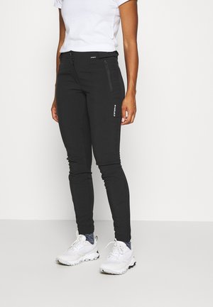 Person iført sorte bukser med lynlås, hvide sneakers med blå sokker og en hvid kortærmet skjorte, stående mod en ensartet lys baggrund.