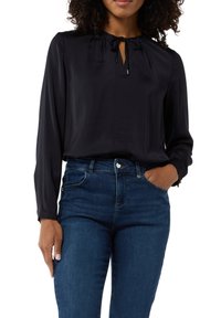 Czarna bluzka z długim rękawem, z zebranym dekoltem i zapięciem na związek, w połączeniu z niebieskimi dżinsami. Gładka tekstura materiału i dopasowany krój.