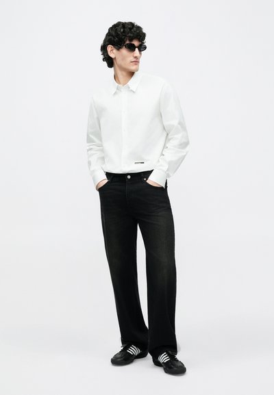 Mand med krøllet hår og solbriller iført en hvid skjorte med knapper, sorte jeans og sorte sneakers med hvide striber, stående med hænderne i lommerne.