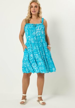 Nainen, joka pukeutuu hihattomaan siniseen, kuvioituun polvipituiseen kesämekkoon ja valkoisiin avoihin varvassandaaleihin, seisoo käsien ollessa mekon taskuissa.