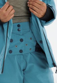 Türkisfarbene Skijacke mit Frontreißverschluss, hohem Kragen und schwarzen Akzenten. Innen dazu passende, hoch taillierte Türkisfarbene Hose mit Knopfdetail.