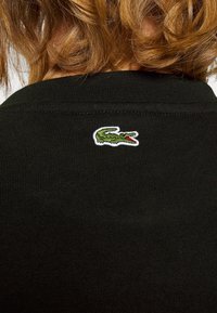 Suéter de punto negro con un pequeño logotipo de cocodrilo verde bordado y boca roja en la parte posterior cerca del cuello.