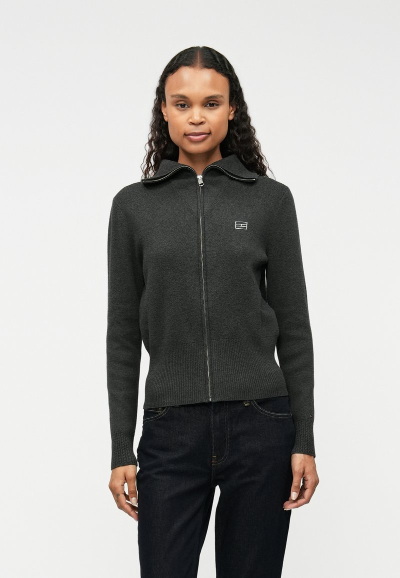 Mørkegrå zip-up genser i mykt materiale, med høy krage, ribbede mansjetter, og en liten logodetalj på venstre bryst.