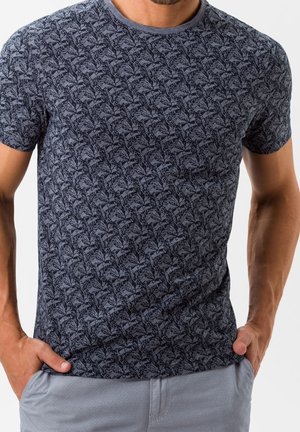 Hombre con camiseta azul marino con estampado, de manga corta, y pantalones gris claro, con las manos en los bolsillos, sobre un fondo liso.