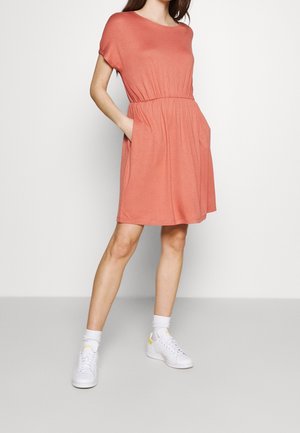 Femme portant une robe ample corail à manches courtes, longueur genou avec des poches, des chaussettes blanches aux chevilles et des baskets blanches, sur un fond uni.