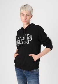 Black cotton hoodie with white "GAP" logo, front pocket, drawstring hood, and ribbed cuffs, paired with blue denim jeans.