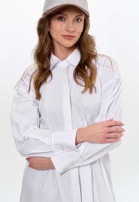 Biała koszula z długim rękawem, gładka w dotyku, z kołnierzykiem. Charakteryzuje się prostym wzorem, z przodem na guziki i guzikami przy mankietach.