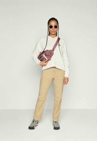 Hvit hettegenser med en liten logo, beige bukser, grå og rosa joggesko, og en brun crossover-veske med stropp. Inkluderer solbriller og flettet hår.