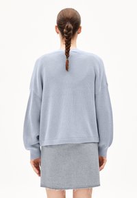 Vaaleansininen neulottu pusero, jossa on väljistynyt malli ja resorikuvio, yhdistetty harmaaseen huopaan minihameeseen, jonka reunat ovat rispaantuneet.