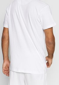 Hombre vestido con una camiseta blanca de manga corta y pantalones cortos blancos, visto desde atrás con los brazos relajados a los lados.