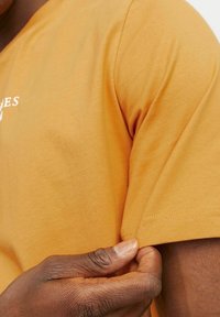 Majica s kratkimi rokavi, okroglim izrezom v gorčično rumeni barvi iz bombaža, ki prikazuje mehak videz in šivani rob, z roko, ki drži rokav.