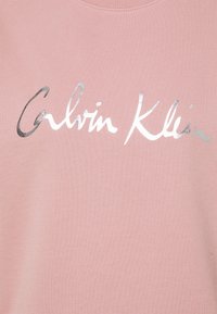 Felpa rosa con testo in argento metallico "Calvin Klein" in font corsivo, tessuto morbido e scollatura a girocollo.