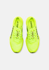 Neon gul atletik sko med mesh-overdel, sorte accenter og flade snørebånd. Bemærkelsesværdig branding på siden og tungebåndet. Letvægtsdesign.