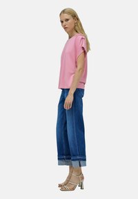 Mujer viste una blusa rosa de manga corta, jeans anchos con puños de color azul, y sandalias de tacón color beige, de perfil sobre un fondo blanco.