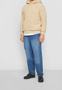Beige, textured fleece hoodie with a kangaroo pocket; paired with loose-fitting blue denim jeans and black sneakers.