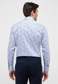Camicia a maniche lunghe con un motivo floreale blu chiaro, realizzata in tessuto dalla consistenza liscia. Indossata con pantaloni scuri che mostrano dettagli con bottoni.