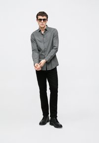 Fekete-fehér kockás hosszú ujjú ing, fekete nadrággal és fekete fűzős csizmával, egyszínű fehér háttér előtt állva.