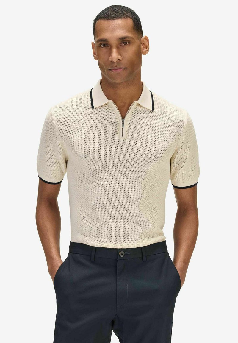 Homme portant un polo en maille beige à manches courtes avec bordure noire et un pantalon bleu marine, debout les mains dans les poches devant un fond uni.