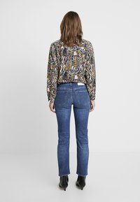 Vrouw met schouderlang bruin haar, gekleed in een kleurrijk geblokt blouse, blauwe jeans en zwarte enkellaarsjes met hak, staand met de rug naar de kijker.
