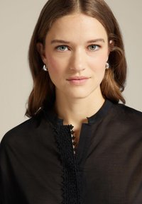 Blusa nera con collo alto, caratterizzata da una bordatura in pizzo sul davanti, tessuto dalla texture liscia e una leggera lucentezza.