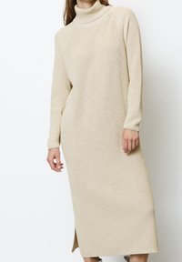 Femme portant une longue robe en maille beige à col roulé, manches longues et fente latérale, debout devant un fond blanc.