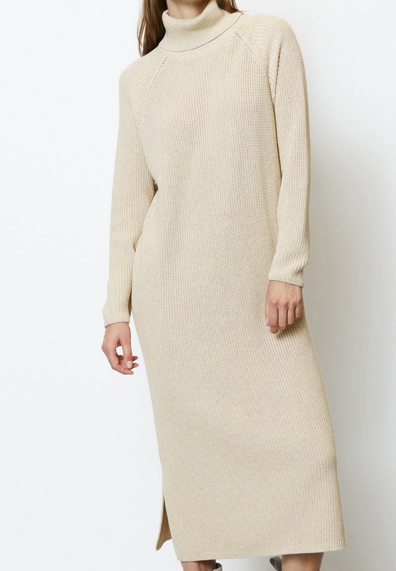 Femme portant une longue robe en maille beige à col roulé, manches longues et fente latérale, debout devant un fond blanc.