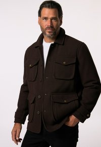 Chaqueta de forro polar marrón con un cuello tipo solapa, dos bolsillos en el pecho, dos bolsillos laterales y cierres de botón. Pantalones negros llevados debajo.
