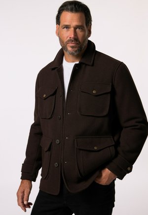 Veste en polaire marron avec un col, deux poches poitrine, deux poches latérales et des fermetures à boutons. Pantalons noirs portés en dessous.