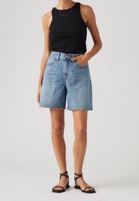 Pantalones cortos de mezclilla azul claro con un corte holgado, cintura alta y diseño de cinco bolsillos. Combinados con una camiseta de tirantes negra acanalada y sandalias negras con tiras.