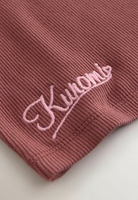 Tissu côtelé rose poussiéreux avec texte en cursive brodé rose « Kuwalla » près de l'ourlet, montrant une texture et des coutures détaillées.