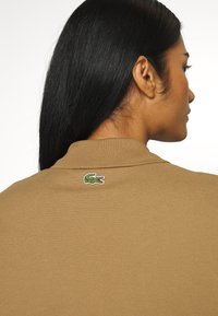 Ruskea polo-paita, jossa on teksturoitu kangas ja vihreä krokotiililogo takakauluksessa. Minimalistinen muotoilu klassisella istuvuudella.