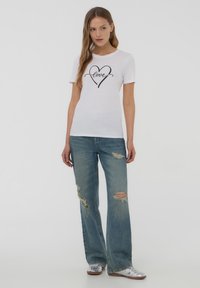 T-shirt bianca a maniche corte con grafica di cuore nero e testo "love", abbinata a jeans strappati blu sbiaditi e sneakers argento.