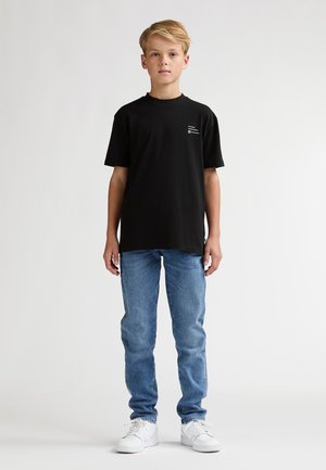 T-shirt en coton noir avec col rond, associé à un jean en denim bleu. Le t-shirt présente un petit logo imprimé sur la poitrine. Baskets blanches.