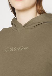 Sudadera con capucha de color verde oliva hecha de material suave, que presenta un sutil logo en gris claro de "Calvin Klein" en el pecho.