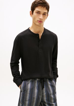 Mladi moški s kratkimi rjavimi lasmi, oblečen v črno henley majico z dolgimi rokavi in modre hlače z vzorcem ter z vrvico, roke v žepih.