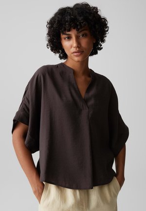 Frau mit lockigem Haar, die eine lockere dunkelbraune kurzärmelige Bluse und hellbeige Hose trägt, mit den Händen in den Taschen, vor einem schlichten Hintergrund stehend.