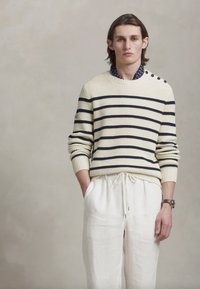 Pull en tricot crème avec des rayures bleu marine, doté de boutons aux épaules. Associé à un pantalon blanc ample, le mannequin a les cheveux longs et un arrière-plan neutre.