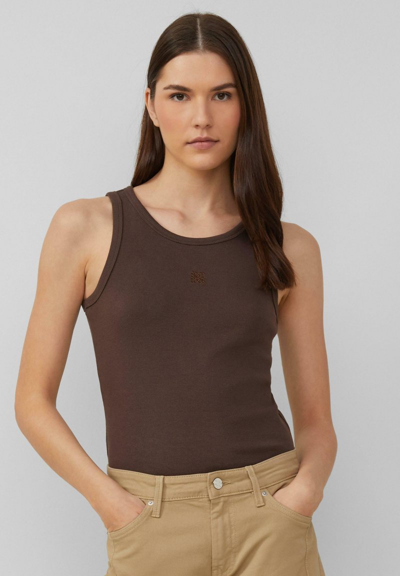 Braunes geripptes Tanktop mit rundem Halsausschnitt, das ein kleines Logo in der Mitte zeigt. Kombiniert mit hellbeigen Hosen, die Hände in den Taschen.