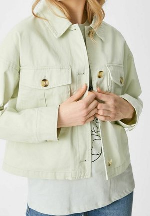 Osoba ubrana w jasnobeżową zapinaną na guziki kurtkę na białą koszulkę z nadrukiem i niebieskie dżinsy, zapinająca guziki kurtki z przodu.