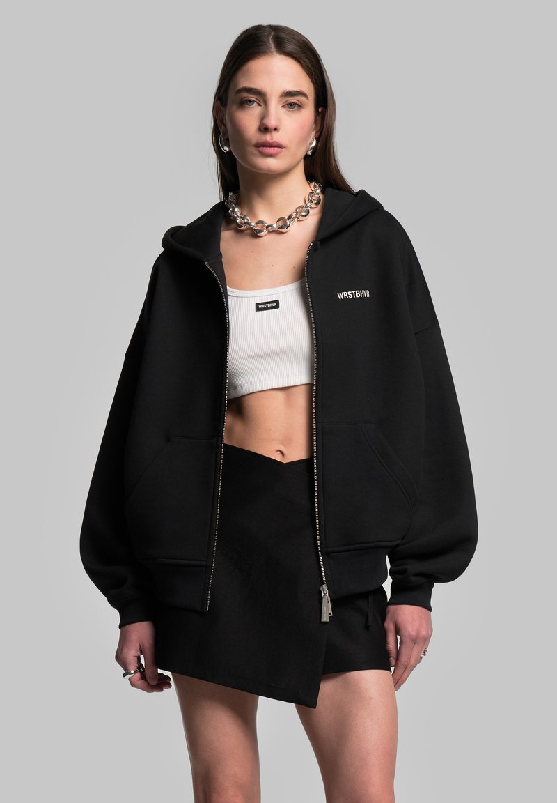 Felpa nera con zip, dalla vestibilità oversized, con polsini a coste e logo sul lato sinistro del petto, abbinata a un crop top bianco fitted e una gonna nera avvolgente.