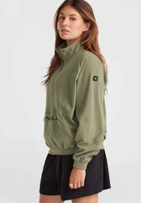 Green fleece pullover with a half-zip collar, elastic cuffs and hem, and a front pocket. Features a logo patch on the left sleeve. Black shorts.