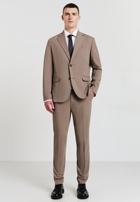 Brauner maßgeschneiderter Anzug mit einem Ein-Knopf-Jackett und maßgeschneiderten Hosen, kombiniert mit einem weißen Hemd und einer marineblauen Krawatte sowie schwarzen Anzugschuhen.