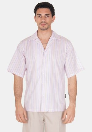 Uomo con camicia rosa chiaro a maniche corte con sottili righe verticali e pantaloni beige, in piedi davanti a uno sfondo bianco.