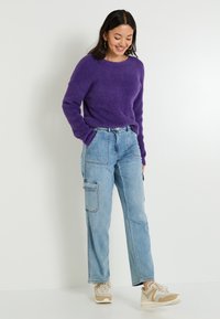 Suéter morado de felpa combinado con jeans cargo de color azul claro que tienen bolsillos laterales y zapatillas blancas con acentos beiges.