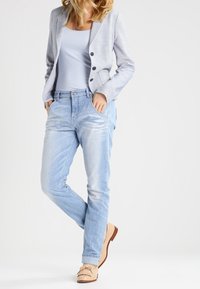 Blazer gris clair avec deux boutons, associé à un débardeur bleu clair et un jean bleu délavé. Les chaussures sont beiges avec détails en franges.