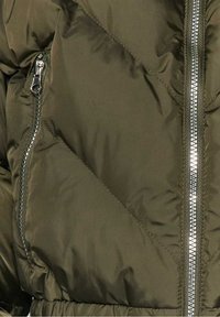 Oddíl olivově zelené péřové bundy se šikmým prošitým vzorem, stříbrným zipem a elastickým pasem. Hladká saténová textura.