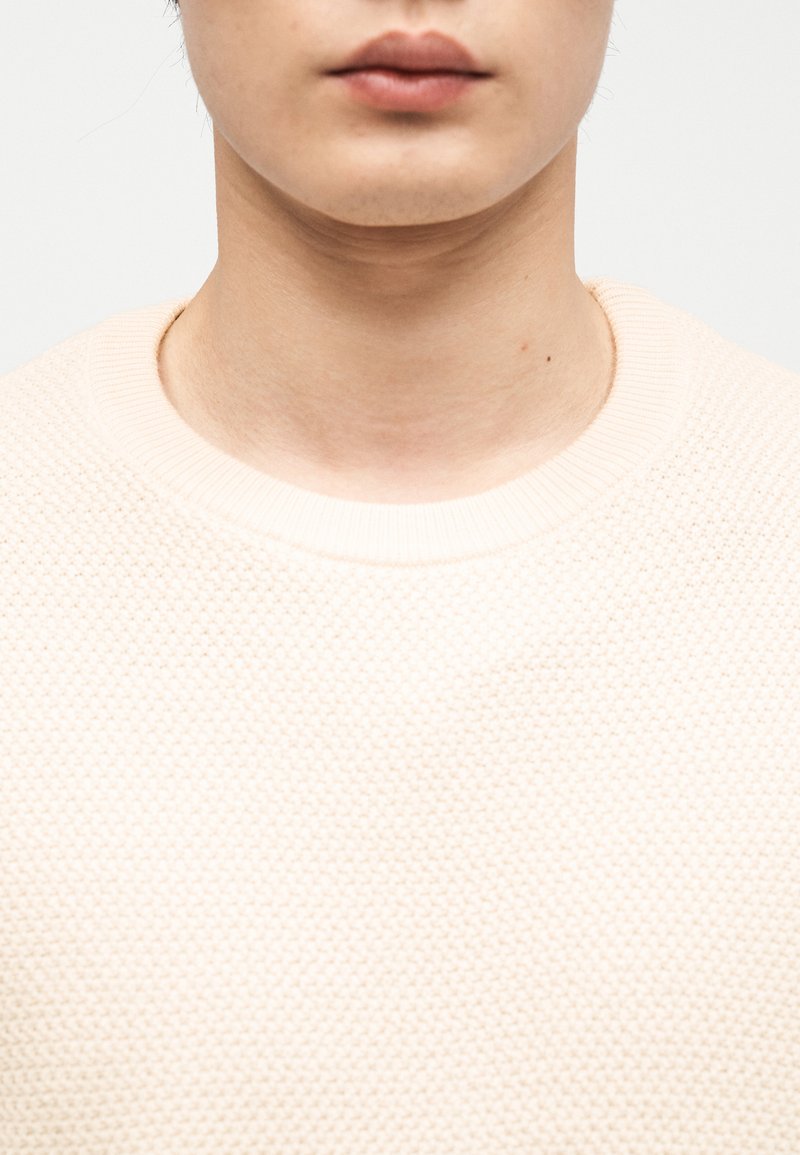 Close-up of a person’s lower face and neck wearing a cream-colored textured knit sweater against a plain light background.