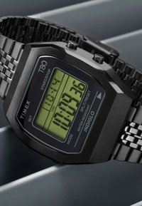 Montre numérique Timex T80 avec un boîtier octogonal noir, un affichage LCD vert et un bracelet en métal avec un design à trois maillons. Comprend des boutons pour le réglage.